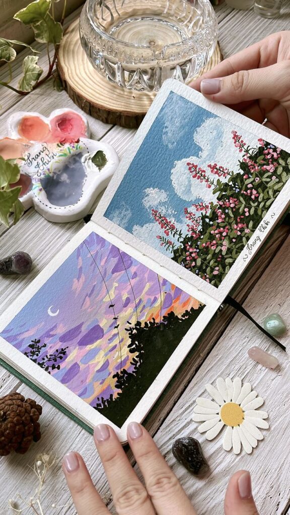 Hands holding a sketchbook with colorful landscape paintings, surrounded by art supplies and crystals on a table.