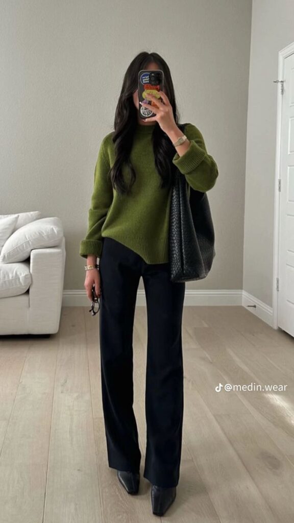 Woman in green sweater and black pants, mirror selfie in stylish living room setting.