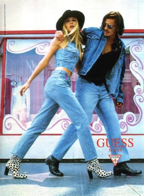 Fashionable duo in denim outfits, striking a pose outside a retro storefront, promoting Guess jeans.