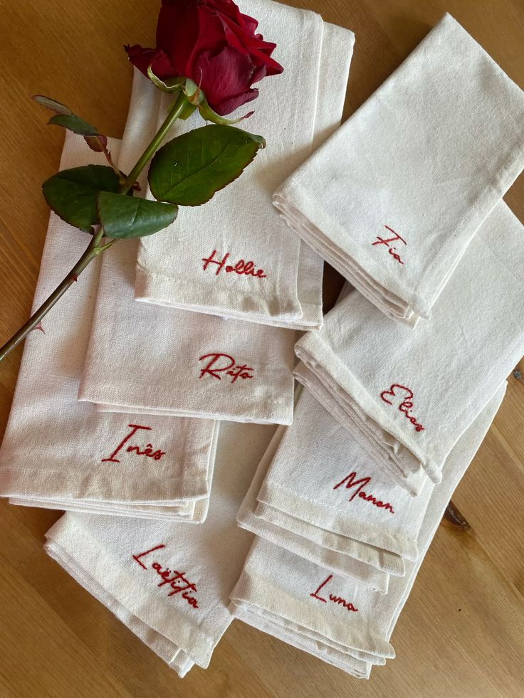 Personalized napkins with names in red embroidery beside a red rose on a wooden table.