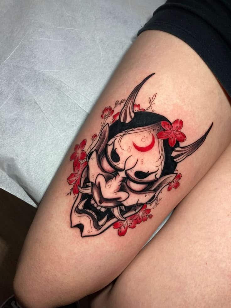 Tattoo of oni mask with red flowers on thigh, showcasing intricate design and bold colors.