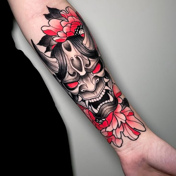 Forearm tattoo of a traditional Japanese oni mask with red eyes, surrounded by vibrant pink flowers.