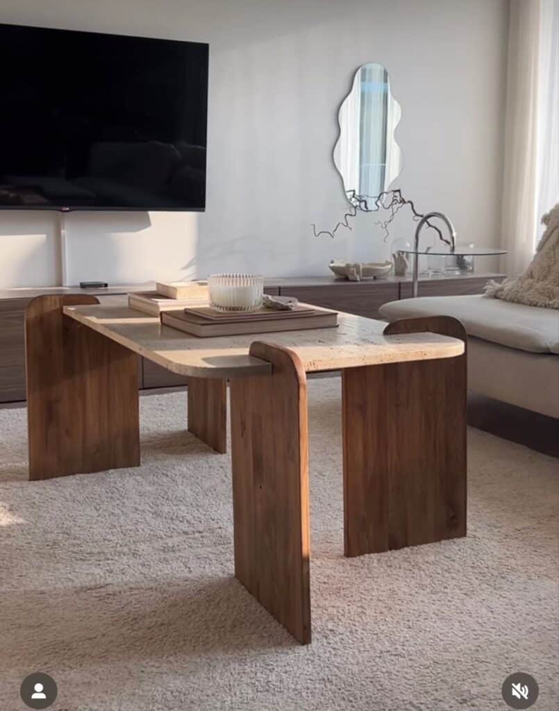 Modern living room with wooden coffee table, abstract mirror, wall-mounted TV, and cozy decor on a beige carpet.