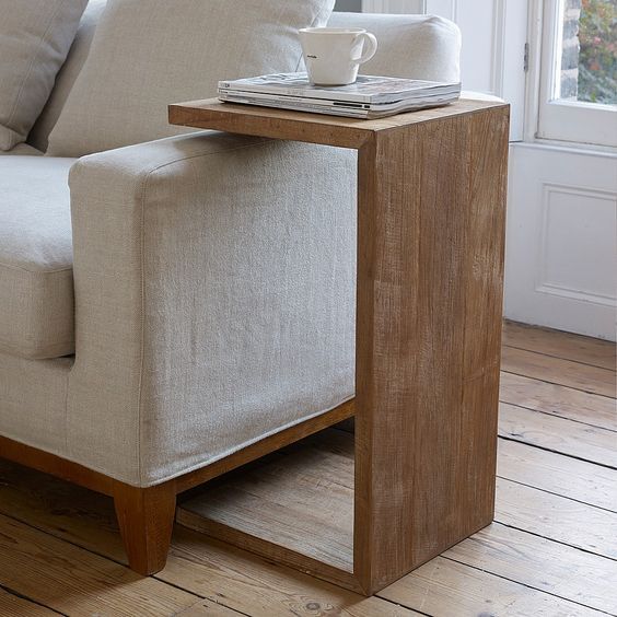 Wooden C-shaped side table with a cup and magazine on a beige sofa, placed on a wooden floor near a window.