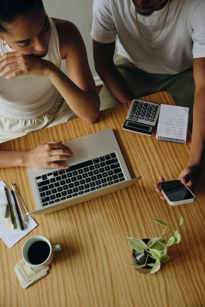 Two people using a laptop, phone, and calculator at a wooden table with coffee and plant, focused on work tasks.