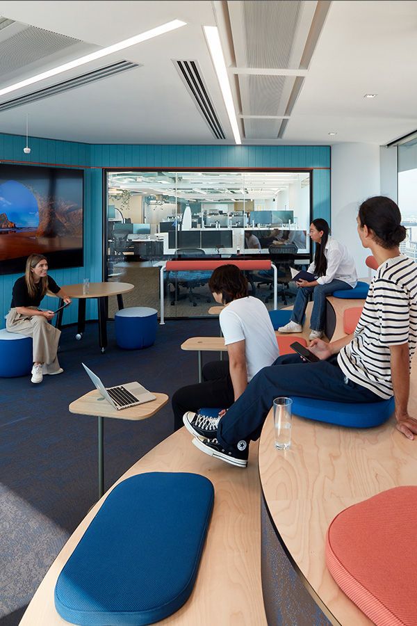 Modern office meeting space with coworkers seated on tiered benches, discussing near a screen and open-plan office.