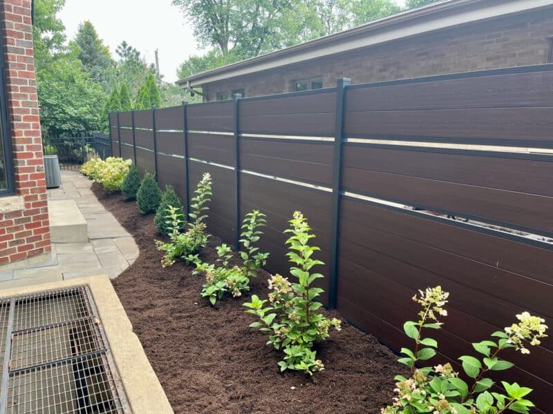 Modern backyard with brown privacy fence, freshly planted shrubs, and lush landscaping along a stone pathway.