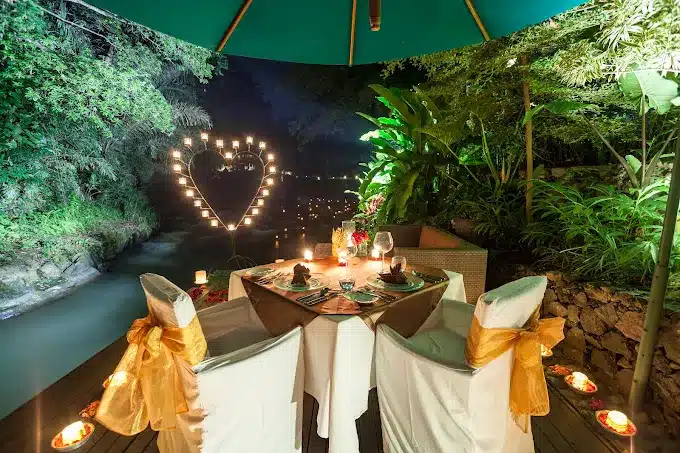 Romantic outdoor dinner setup by a river with candlelit heart decor and lush greenery under a canopy at night.