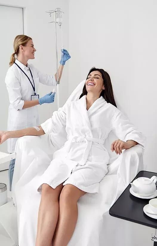 Woman in a white robe receiving an IV drip in a spa setting, attended by a smiling nurse.