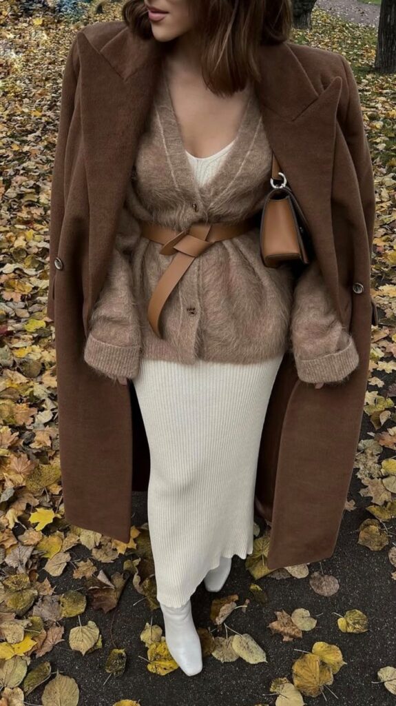 Stylish autumn outfit with brown coat, fuzzy sweater, white skirt, and boots amid fallen leaves.