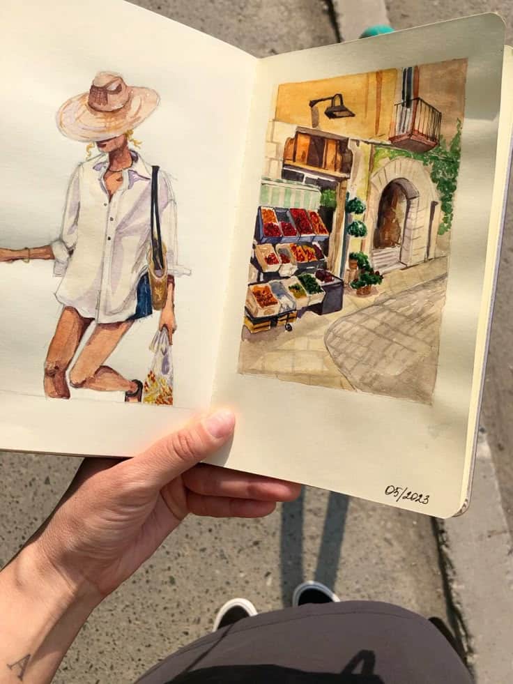 Open sketchbook showing watercolor paintings of a stylish woman and a vibrant market scene, held by a person.