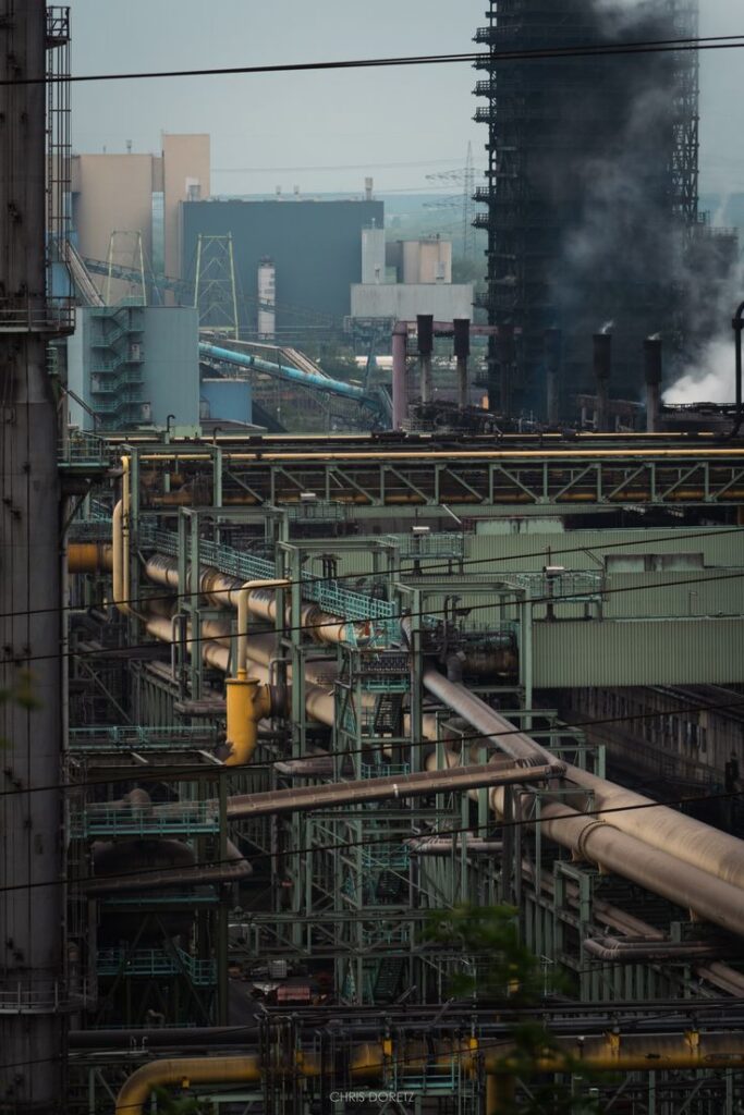 Industrial factory landscape with metal structures, large pipes, and smoke against an urban backdrop.