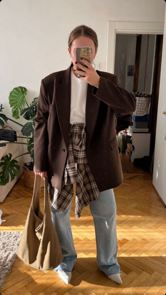 Person in casual outfit with brown blazer, plaid shirt, and jeans, holding a bag in a stylish room.