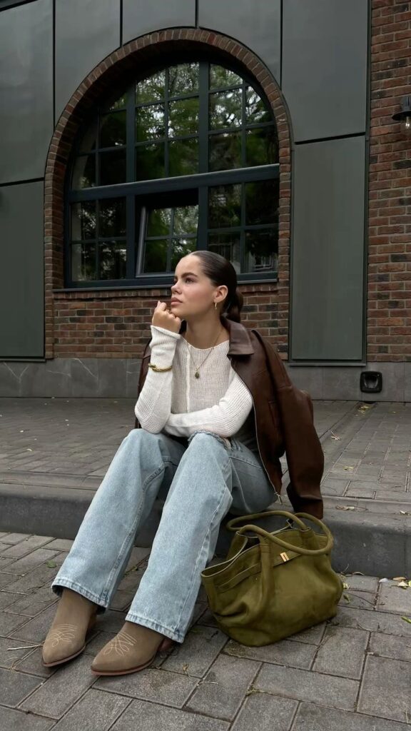 Young woman in casual outfit sitting on steps, wearing a leather jacket and jeans, with a green bag on the ground.
