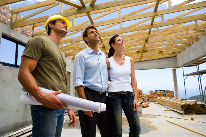 Construction workers and architects inspect a building site, examining the wooden roof framework under a clear sky.