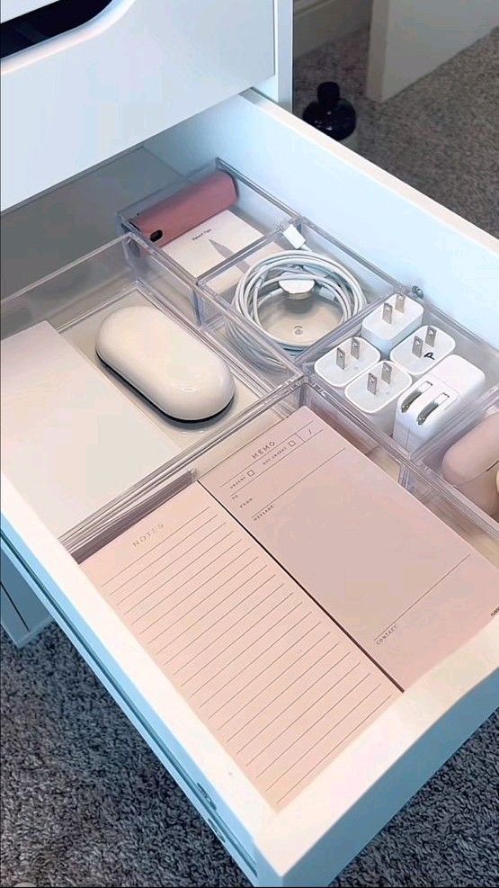 Organized drawer with stationery, cables, adapters, and notepads in clear compartments on a gray-carpeted floor.