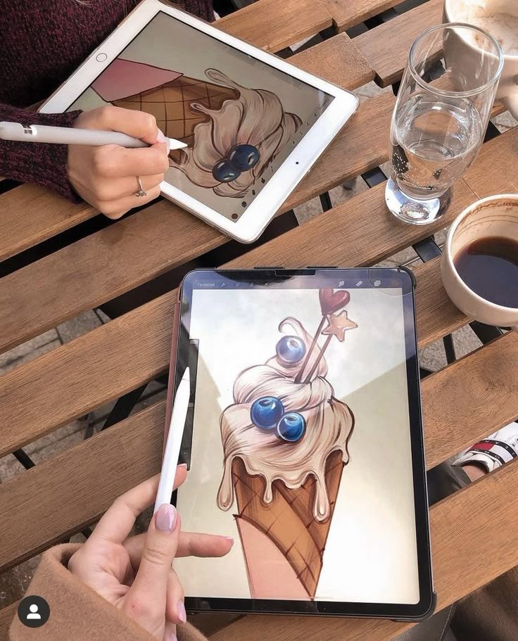 Two people digitally illustrating ice cream cones on tablets, with coffee and water on a wooden table.