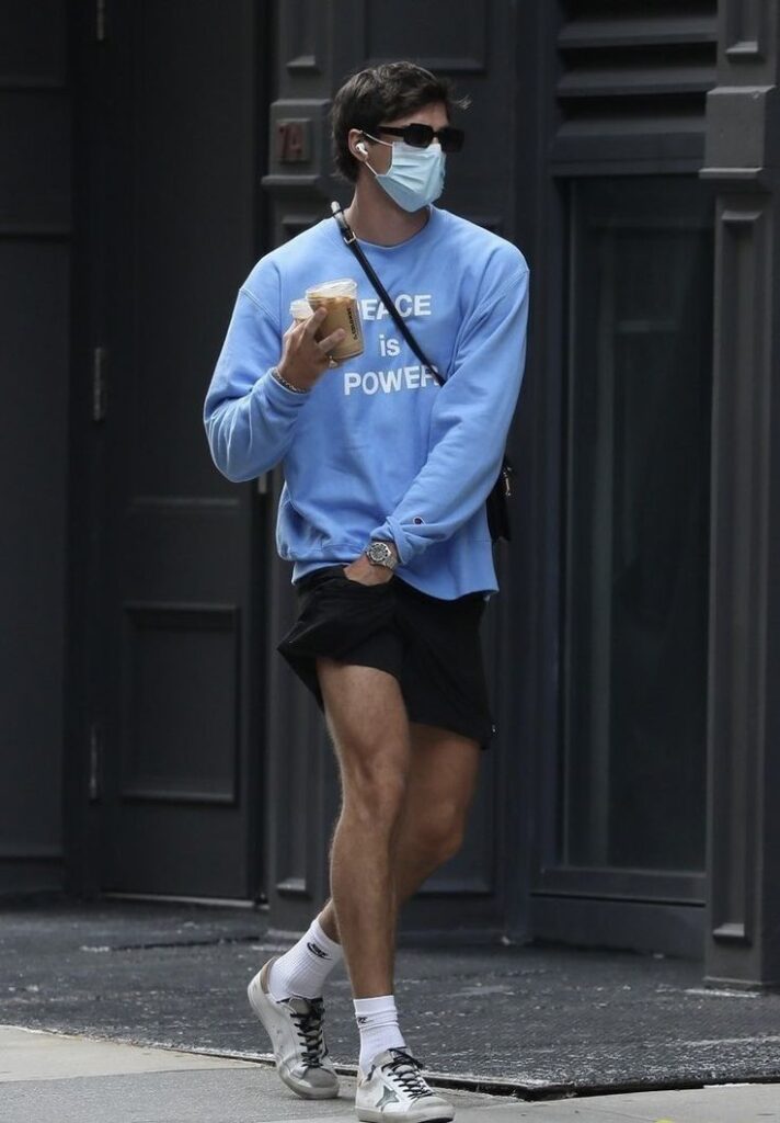 Man in blue Peace is Power sweatshirt walking with iced coffee, mask, and sunglasses on a city street.