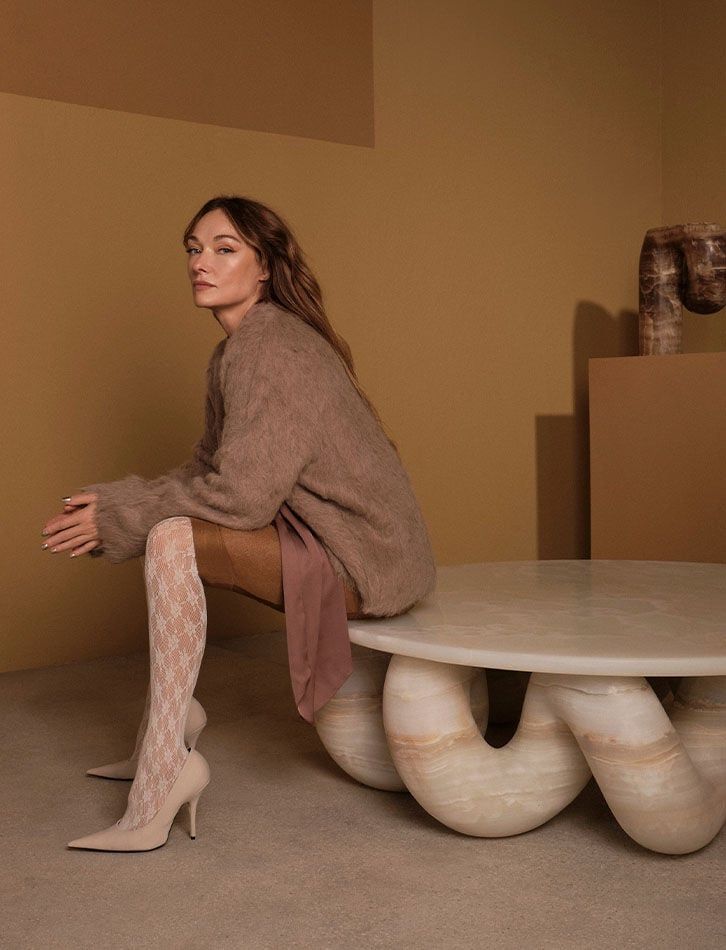 Woman in chic outfit sitting on marble table in beige room, modern style, earthy tones, textured tights, high heels.
