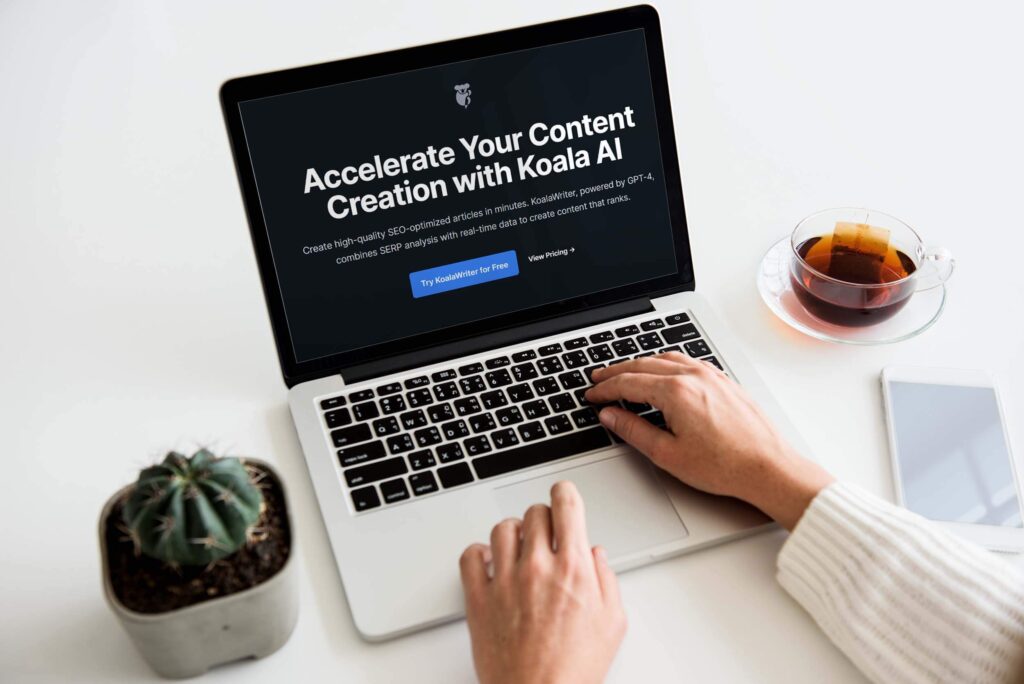 Person using laptop with Koala AI for content creation. Nearby: tea, smartphone, and cactus.