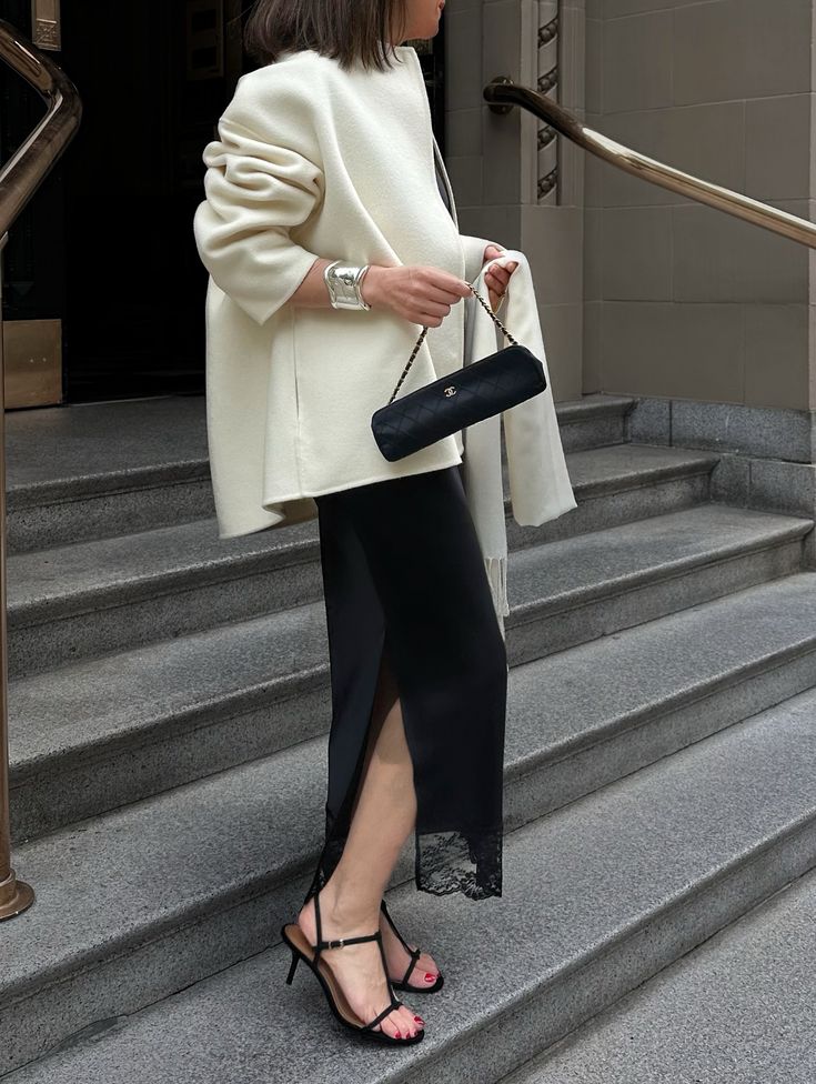 Stylish woman in white coat and black dress with heels, holding a chic clutch on city steps.