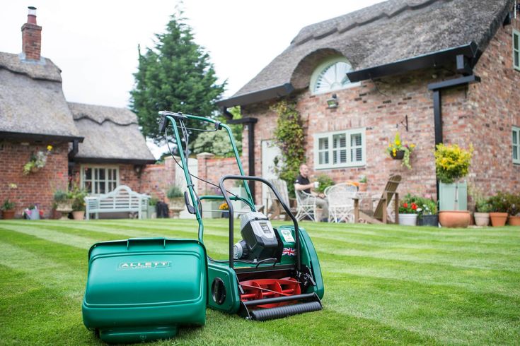 Green lawnmower on neatly trimmed grass in front of charming brick cottage with patio furniture.