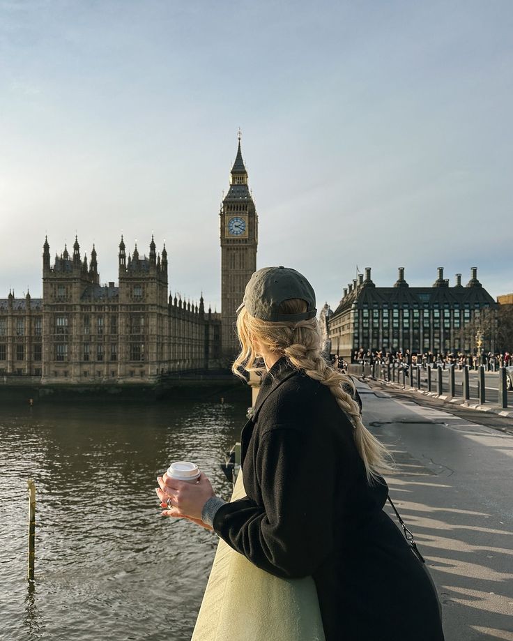 Woman with coffee gazes at Big Ben and the Houses of Parliament in London, capturing scenic river view.