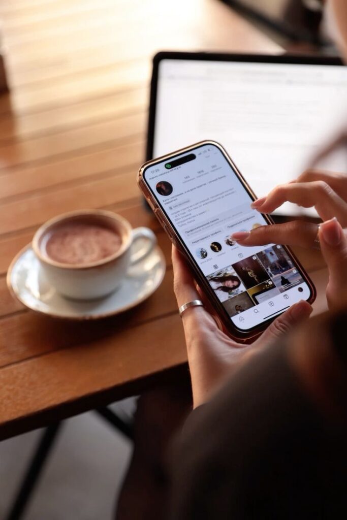 Person browsing social media on smartphone at café with coffee and laptop.