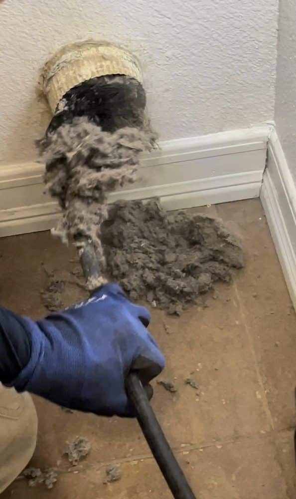 Cleaning dryer vent with tool, removing lint buildup for improved efficiency and safety. Blue glove in action.