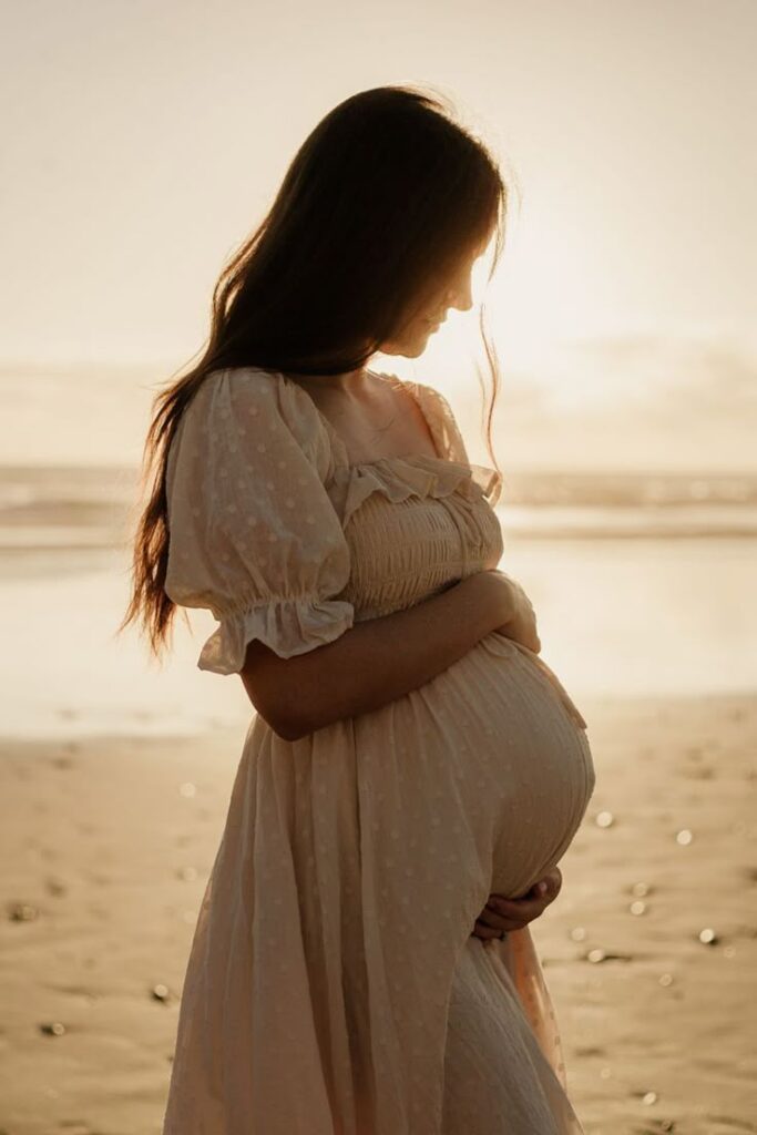 Pregnant woman in a white dress standing on a serene beach at sunset, embracing her belly.