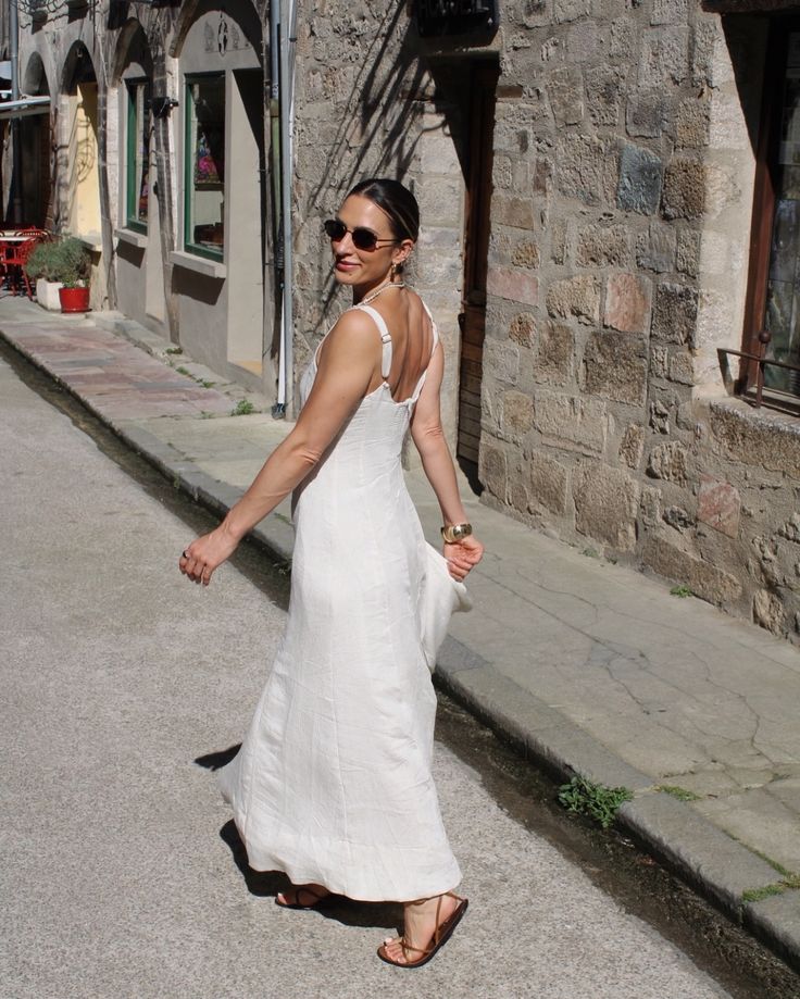 Woman in a white sundress walks down a charming cobblestone street, smiling in the sunshine.