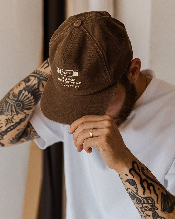 Man wearing a brown cap and white shirt adjusts his hat, showcasing tattoos on his arm.
