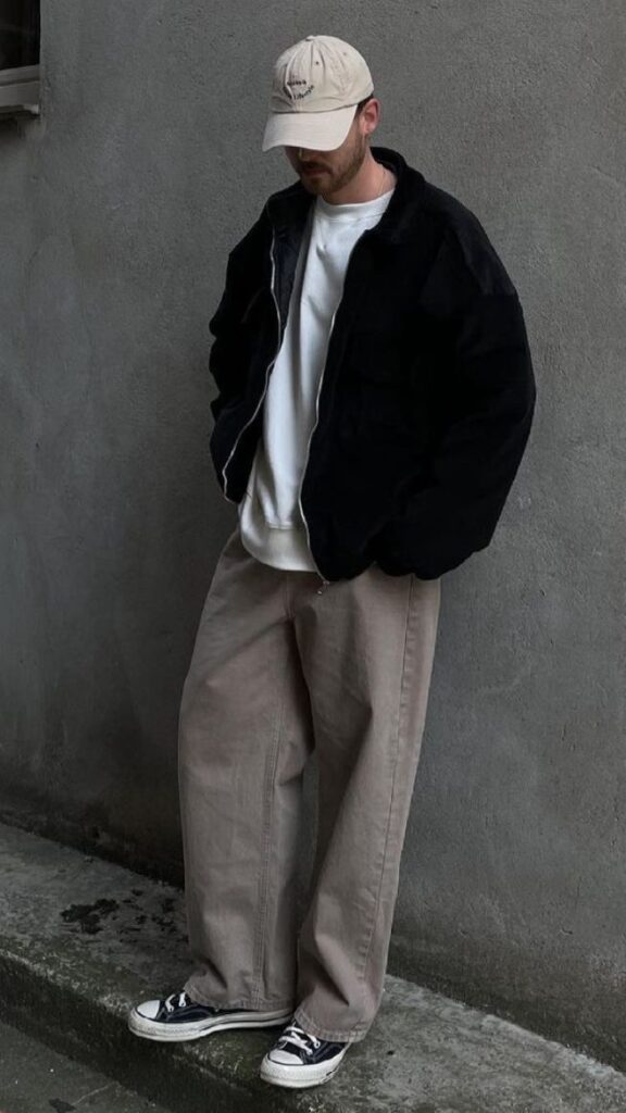 Man in casual streetwear: black jacket, beige pants, white sneakers, cap. Urban fashion against a gray wall.