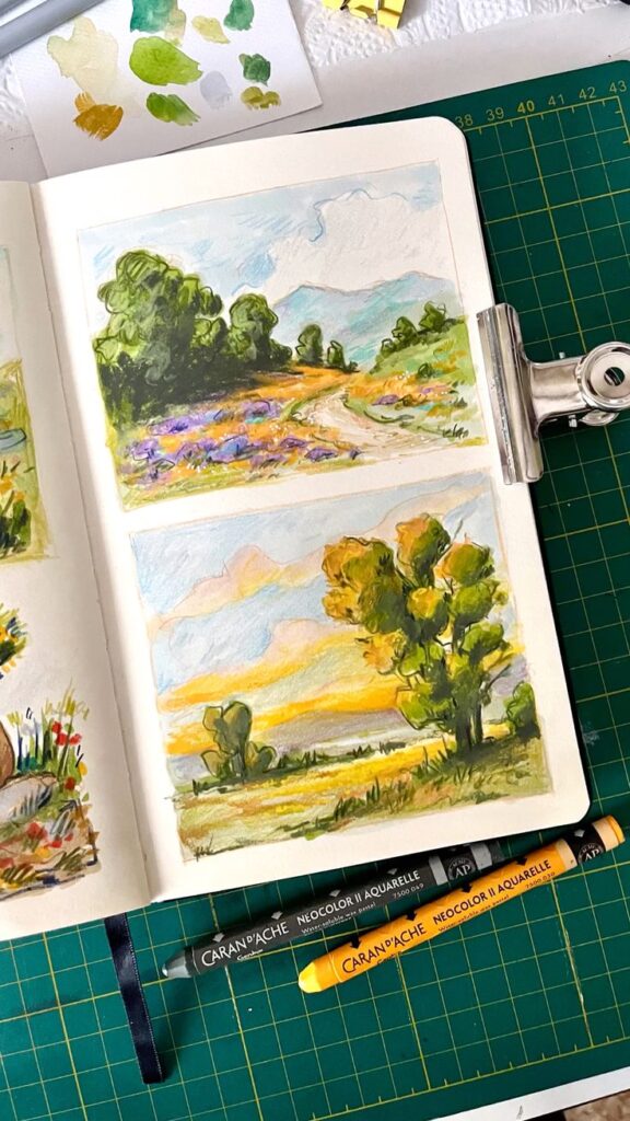 Sketchbook with colorful landscape drawings and Caran d'Ache watercolor pastels on a cutting mat.