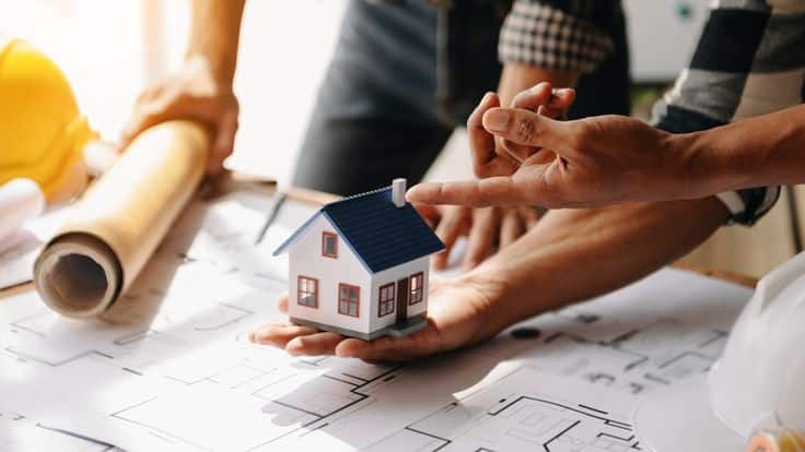 Hands pointing at a miniature house model on blueprints, depicting home design and construction planning.