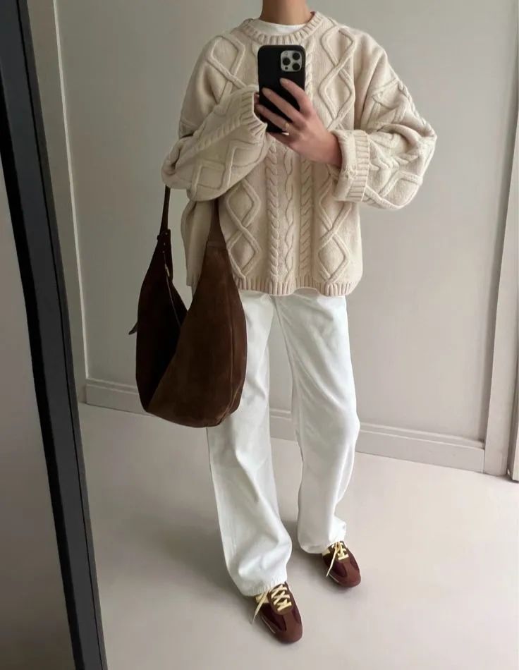 Person wearing cable knit sweater and white pants with brown bag and shoes, capturing mirror selfie indoors.