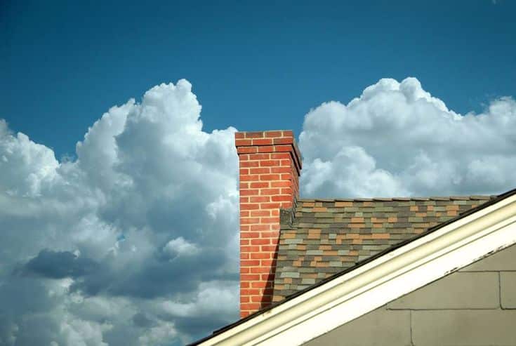 Brick chimney on a roof against a backdrop of bright blue sky and fluffy clouds.