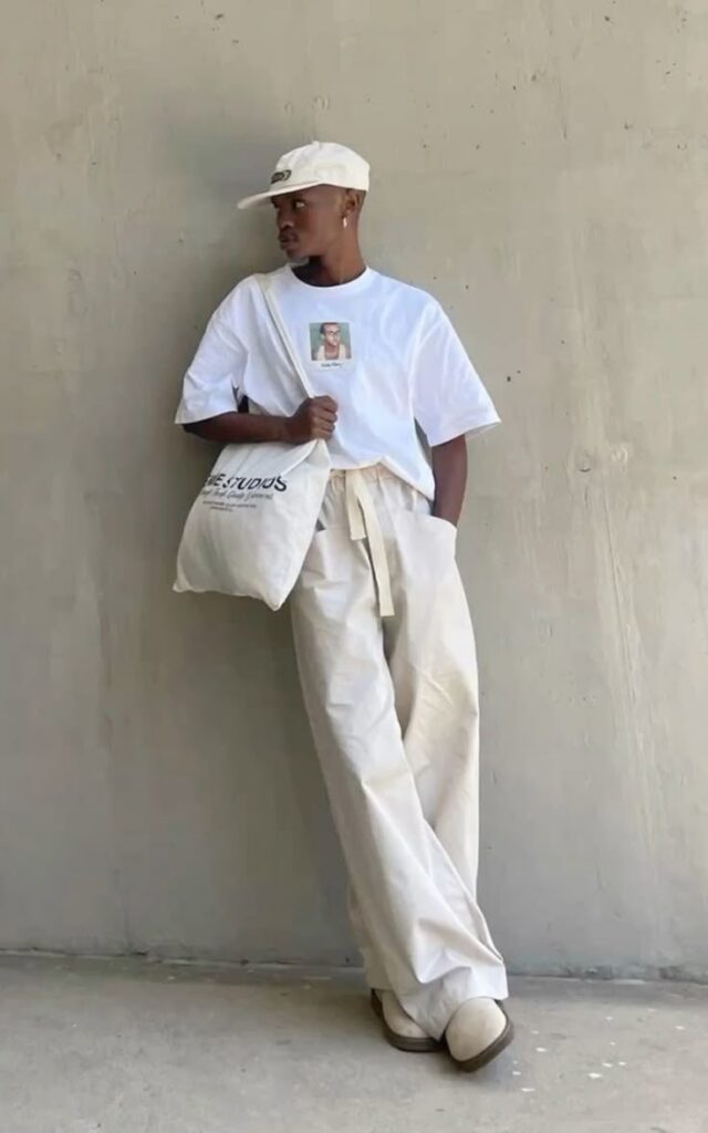 Fashionable person in casual white outfit with tote bag, leaning against a wall, urban style.