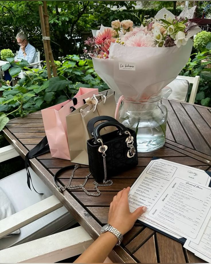 Luxury handbag and bouquet on a wooden table at a café, with a menu and shopping bags.