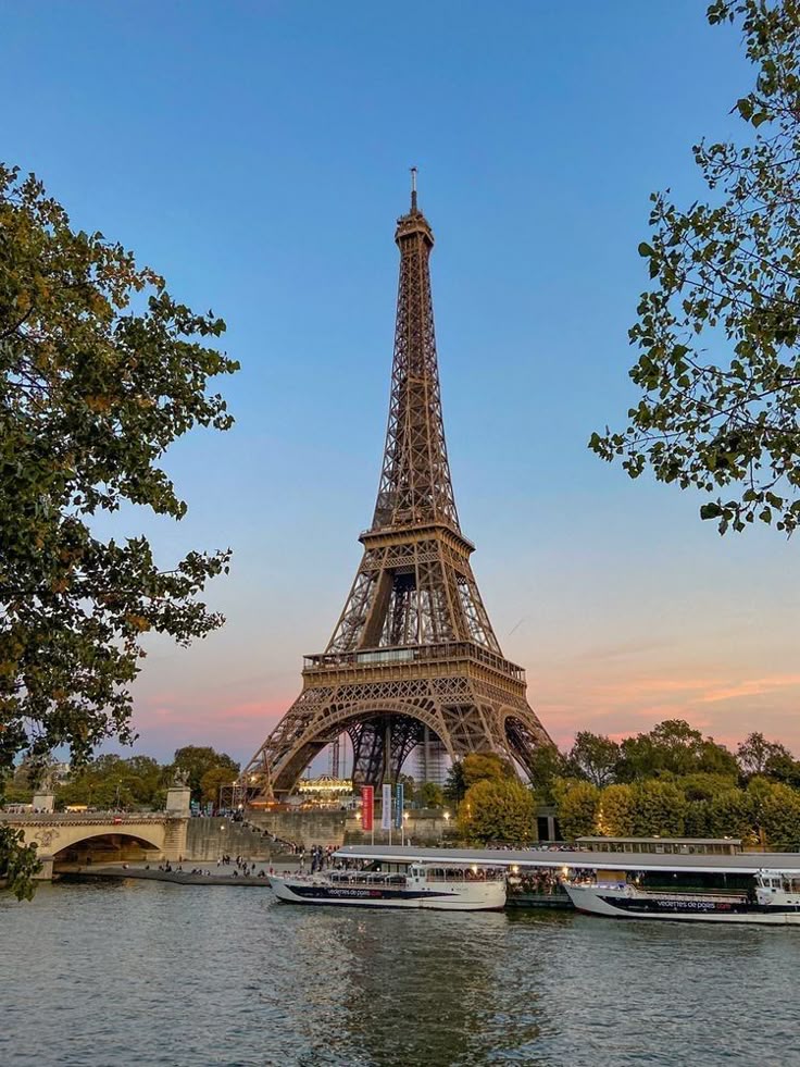 Eiffel Tower at sunset with boats on Seine River and trees, Paris landmark view in vibrant sky backdrop.