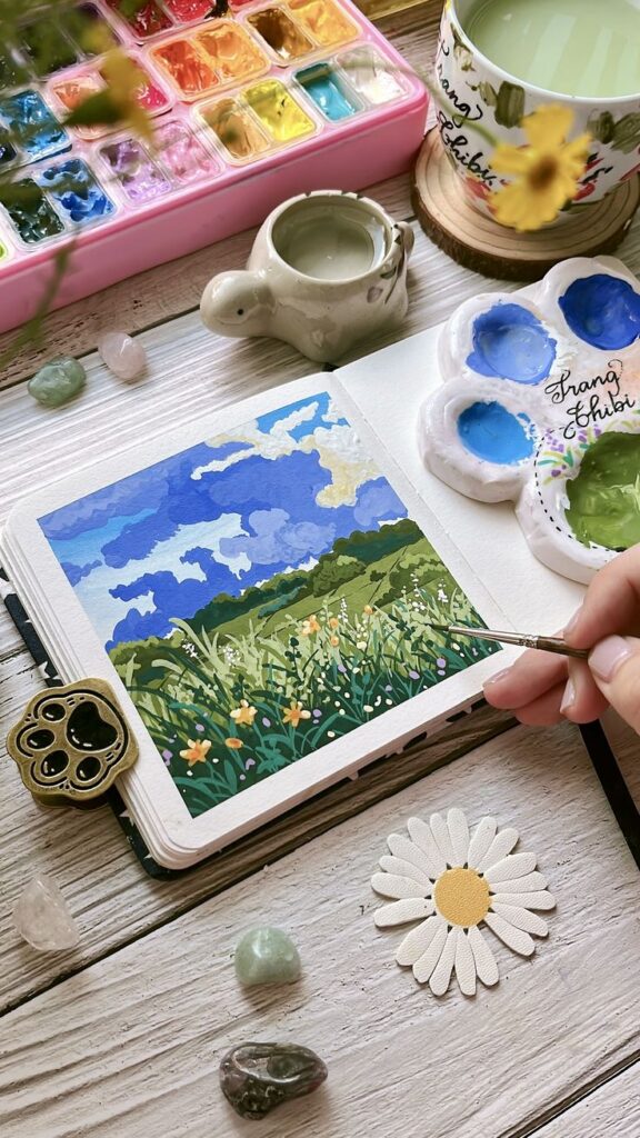 Artist painting a colorful landscape in a sketchbook, surrounded by watercolors and nature-themed items.