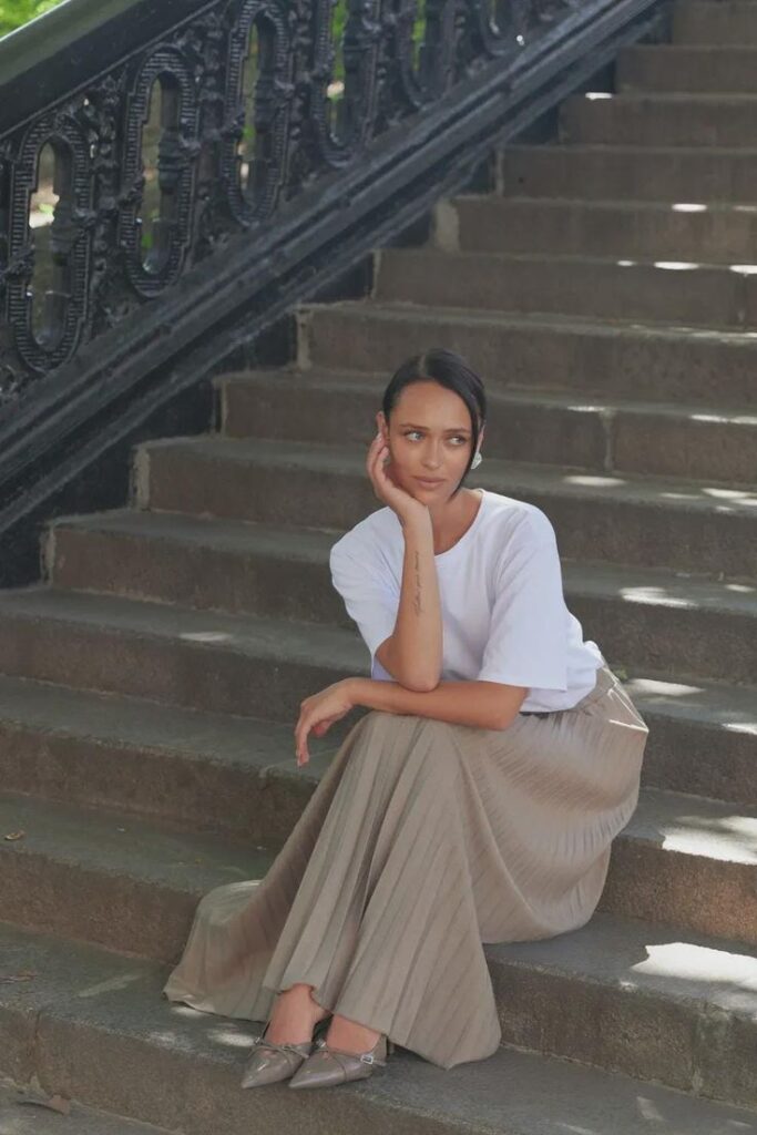 Woman in white top and beige skirt sitting thoughtfully on outdoor stairs.
