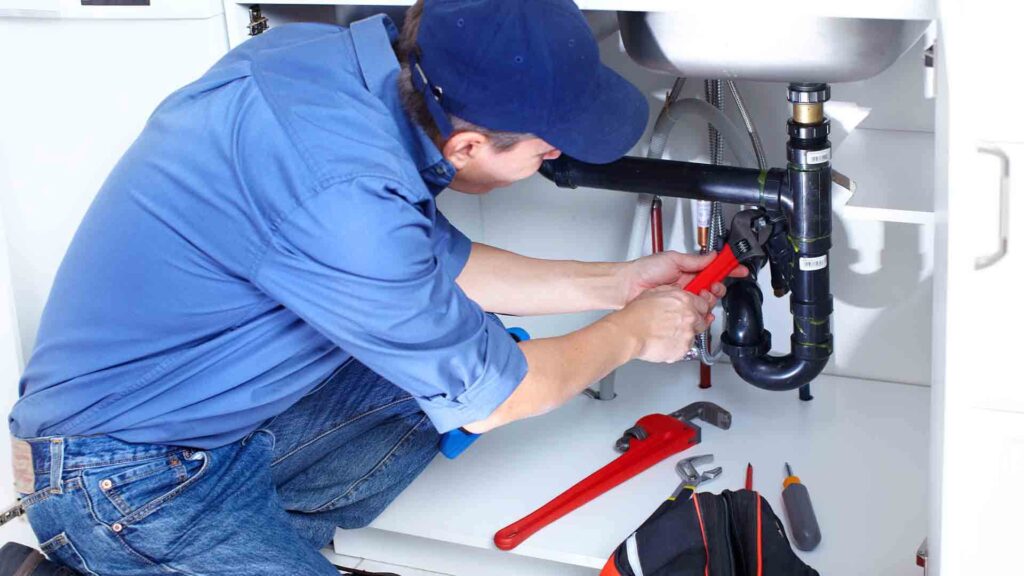 Plumber fixing kitchen sink pipe with wrench, wearing blue uniform and cap. Repair tools visible in foreground.