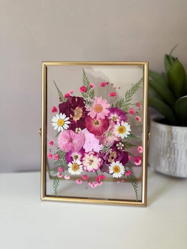 Pressed flowers in a gold frame with pink and white blooms, displayed on a table with a plant in the background.