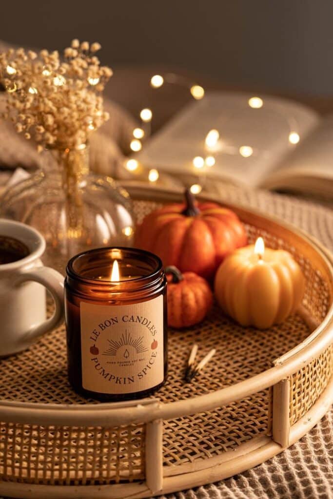 Cozy autumn scene with lit pumpkin spice candle, mini pumpkins, and fairy lights on a woven tray.