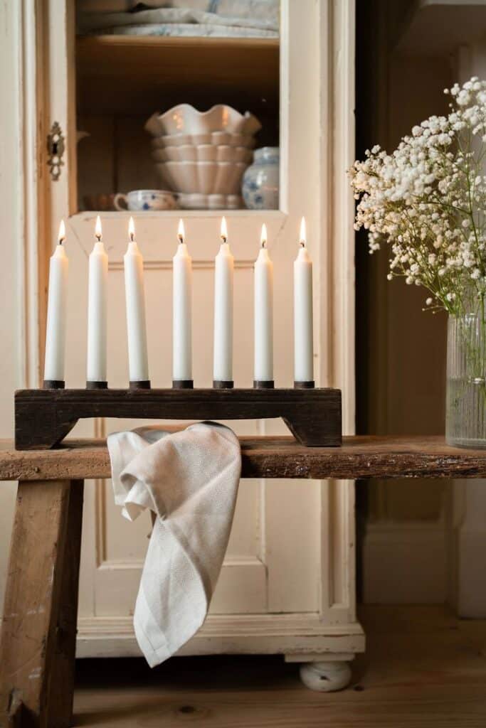 Rustic decor with lit candles on a wooden stand, white cloth, and a vase of flowers in a cozy, vintage setting.