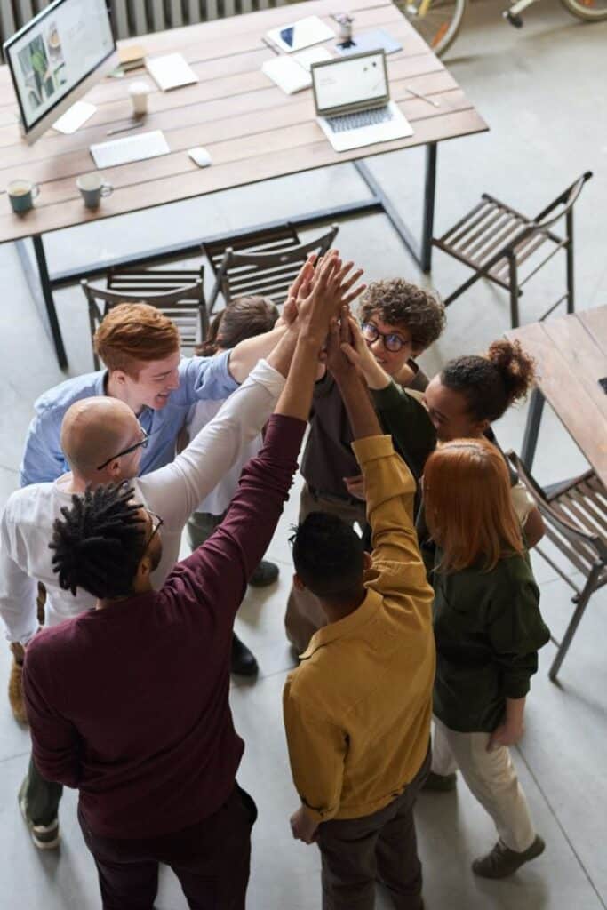 Team celebrating success with a group high-five in office setting. Laptops and coffee cups on wooden table nearby.