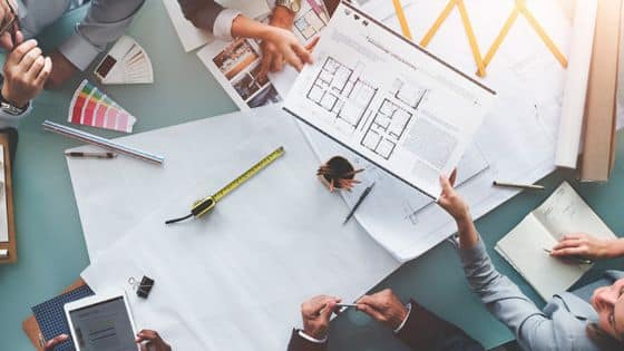 Resilient Advantage Designing and Constructing Facilities Team collaborating on architectural blueprints at a meeting table with plans, tools, and color swatches. | Sky Rye Design Team collaborating on architectural blueprints at a meeting table with plans, tools, and color swatches.