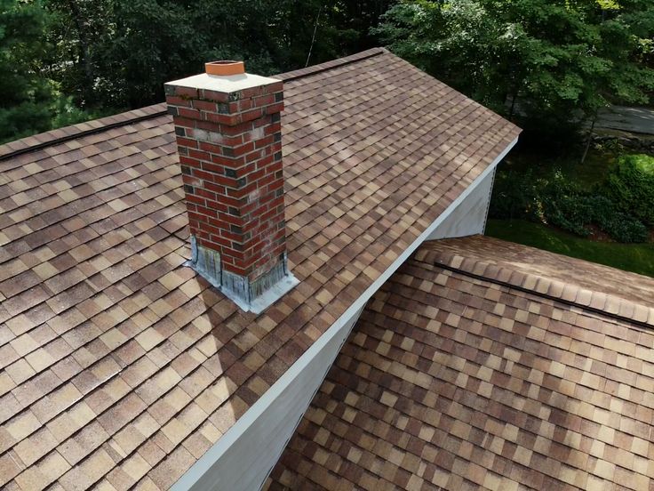 Brown shingle roof with red brick chimney, surrounded by trees and greenery.