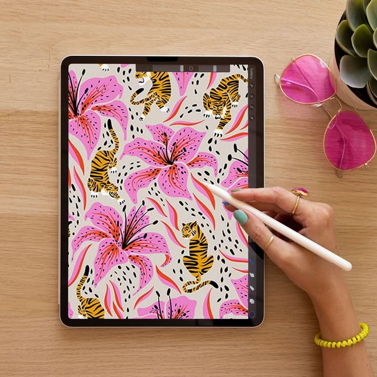 Tablet displaying floral and tiger pattern art, hand holding stylus. Pink sunglasses and plant nearby.